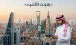 غلاف فوتوغرافي لمدينة سعودية حديثة ورجل يستخدم هاتفاً ذكياً مع رموز متصلة ترمز إلى إنترنت الأشياء (Internet of Things - IoT).
