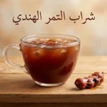 كوب زجاجي شفاف مملوء بشراب التمر الهندي البني المحمر مع قطع ثلج بجانبه لبّ التمر الهندي الخام على طاولة خشبية