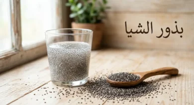 بذور الشيا مبعثرة على سطح خشبي مع ملعقة خشبية وكوب يحتوي على بذور الشيا المنقوعة المتحولة إلى مادة هلامية