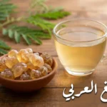 قطع صمغ عربي ذهبية شفافة في وعاء فخاري بجانب كوب ماء مذاب فيه الصمغ العربي على طاولة خشبية مع غصن أكاسيا أخضر في الخلفية ونص "الصمغ العربي"