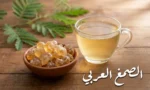 قطع صمغ عربي ذهبية شفافة في وعاء فخاري بجانب كوب ماء مذاب فيه الصمغ العربي على طاولة خشبية مع غصن أكاسيا أخضر في الخلفية ونص "الصمغ العربي"