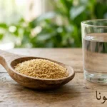 ملعقة خشبية تحتوي على قشور بذور القاطونا الذهبية بجانب كوب ماء زجاجي على سطح خشبي مع كلمة بذور القاطونا