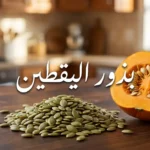 حفنة من بذور اليقطين الخضراء النيئة على سطح خشبي بجوار نصف ثمرة يقطين مفتوحة مع نص عربي يقول بذور اليقطين